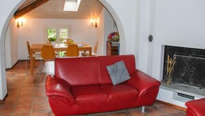 En rød lædersofa med en grå pude i en stue med pejs, en buet døråbning der fører til et spiseområde med bord og stole, og et flisegulv.