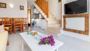 Una sala moderna con un sofá blanco, una mesa de comedor con sillas, una escalera, una zona de cocina y un televisor montado en la pared.