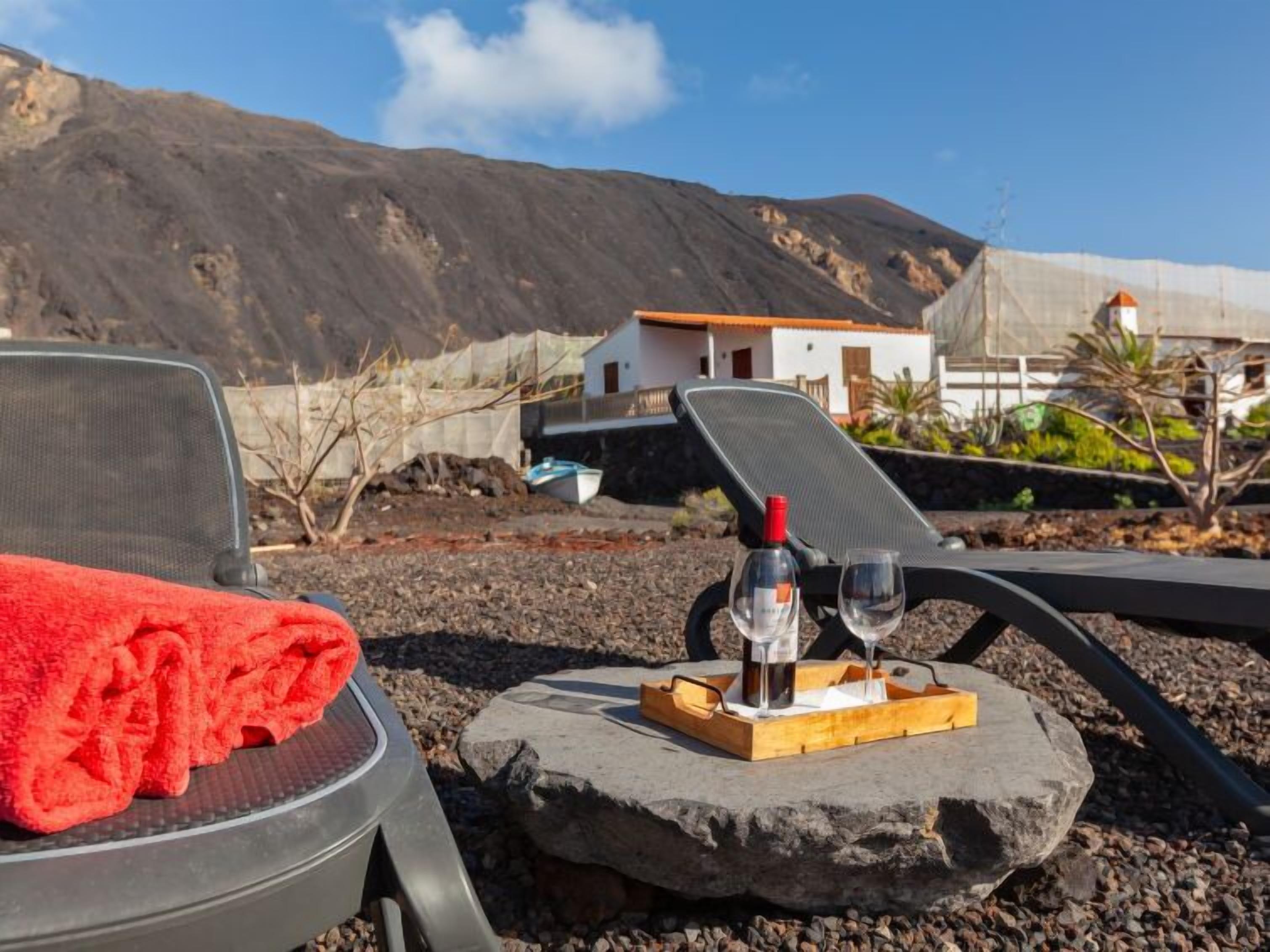 A beach chair with a red towel, a bottle of wine, and two glasses on a tray, set against a rocky beach with a white building and mountains in the background.