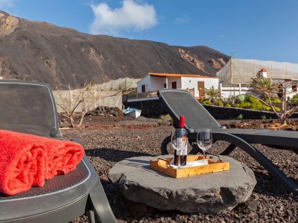 Une chaise de plage avec une serviette rouge, une bouteille de vin et deux verres sur un plateau, le tout placé devant une plage rocheuse, avec un bâtiment blanc et des montagnes en arrière-plan.