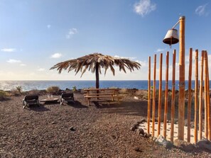 Une plage avec un parasol en chaume, deux chaises, un banc en bois et une clôture en bambou.