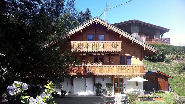 Un chalet de dos pisos con balcón, una pequeña construcción anexa y un jardín cuidado.