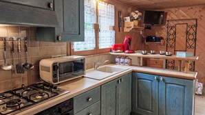 Cocina compacta con estufa a gas, microondas, tostadora y un lavamanos. El techo es de madera y hay una ventana con cortinas de encaje.