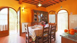 Un comedor con una mesa de madera dispuesta para cuatro personas, rodeada de sillas. Se observa un armario con puertas de vidrio al fondo y un frutero en la encimera.