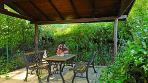 Un patio al aire libre con una mesa de madera y dos sillas, rodeado de abundante vegetación.