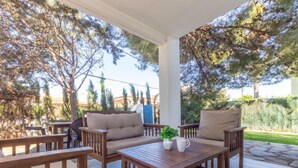 Un espacio de estar al aire libre con techo, mobiliario de madera y una mesa, rodeado de árboles y una vista de un edificio al fondo.