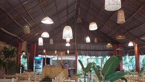 Un restaurante con bancos y mesas de madera, lámparas colgantes tejidas y vistas a zonas verdes al exterior.
