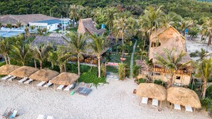 Un complejo turístico en la playa con cabañas de techo de paja, palmeras y tumbonas.