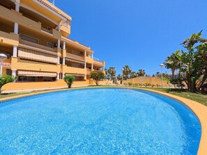 En swimmingpool med krystalklart, blåt vand, omgivet af en gul bygning med flere altaner og palmer.