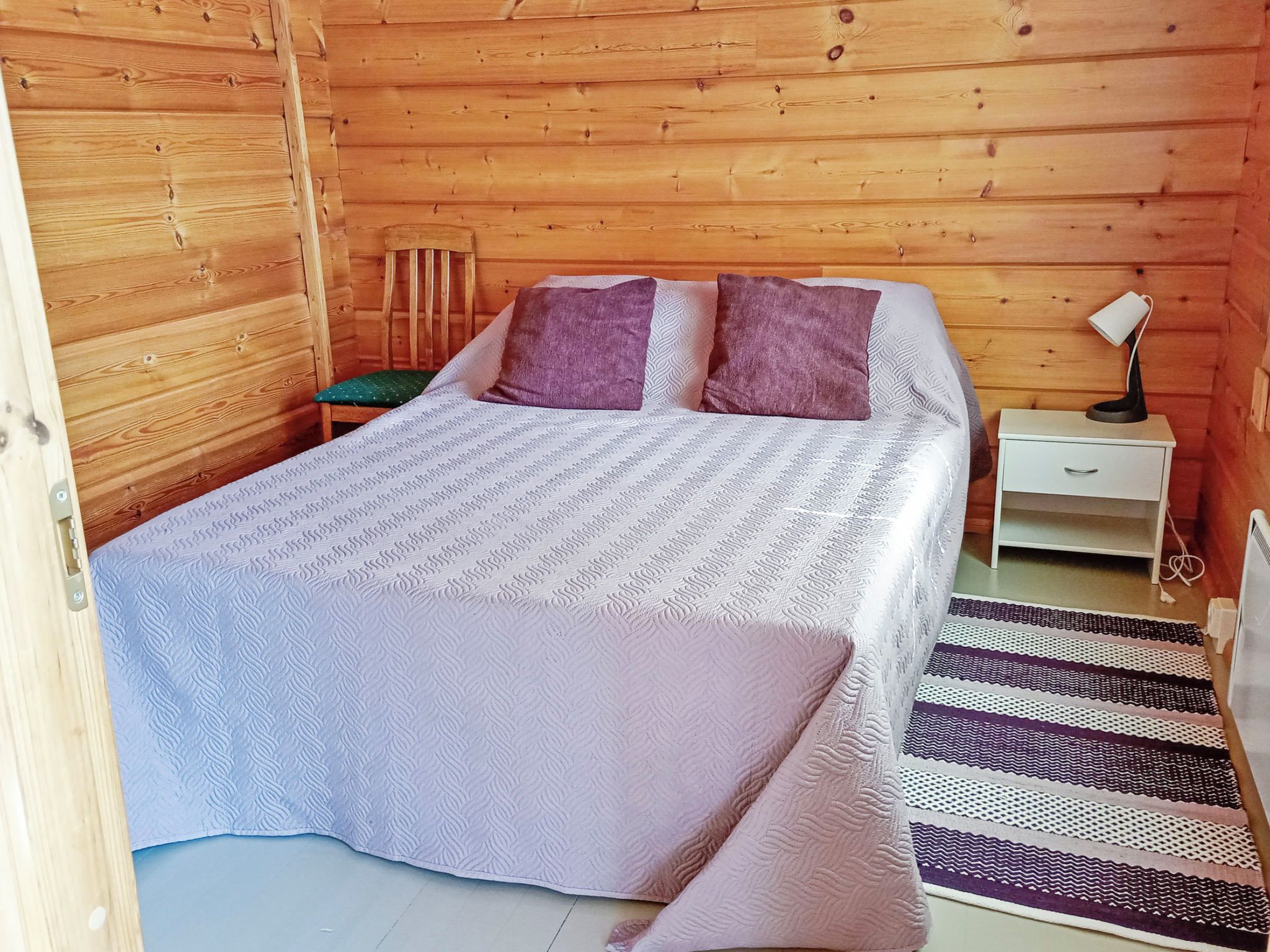 Una habitación de madera con una cama, almohadas, una mesita de noche, una lámpara y una alfombra estampada.