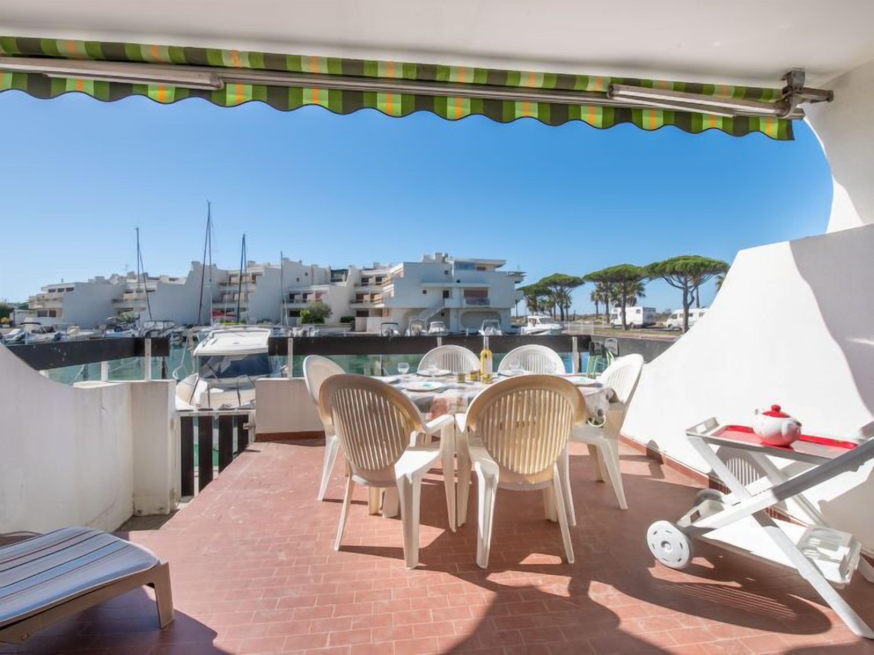 A terrace with a table and chairs, overlooking a marina with boats and buildings.