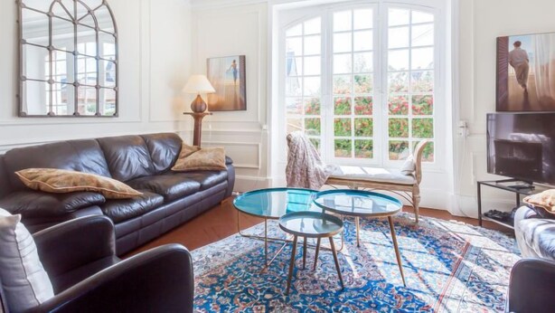 A living room with a black leather sofa, a glass coffee table, a patterned rug, a chandelier, and a large window with a view of greenery. - Le Cyclamen by Interhome (Courseulles)