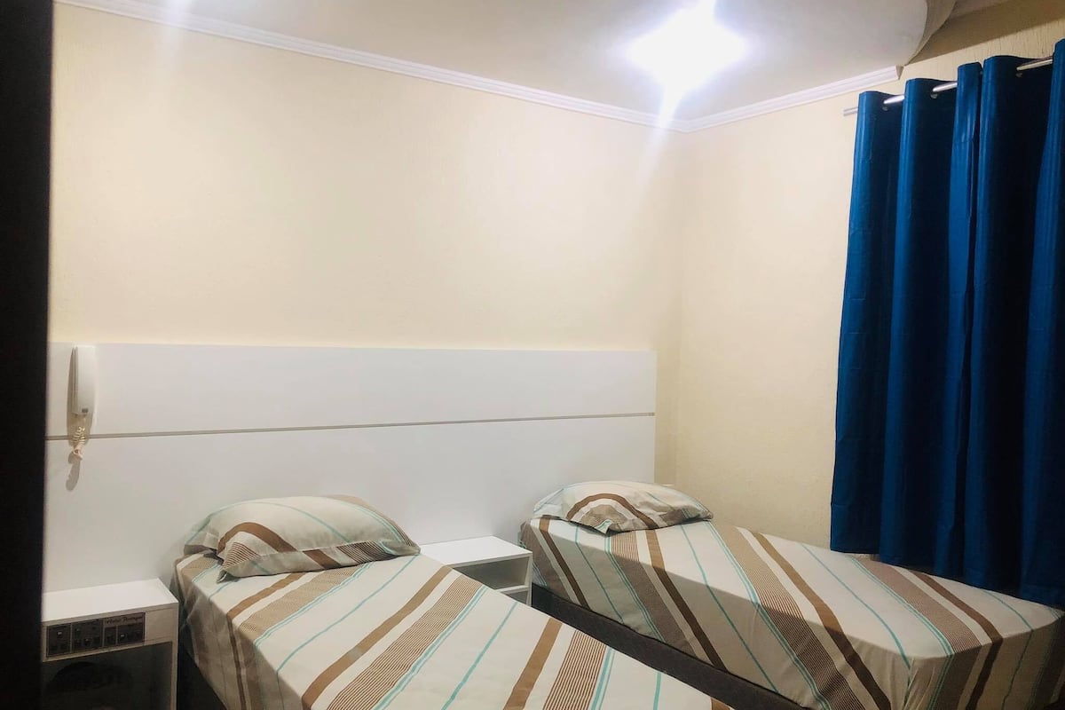 Une petite chambre d’hôtel composée d’une seule pièce, avec deux lits, une table de chevet, un téléphone et un rideau.