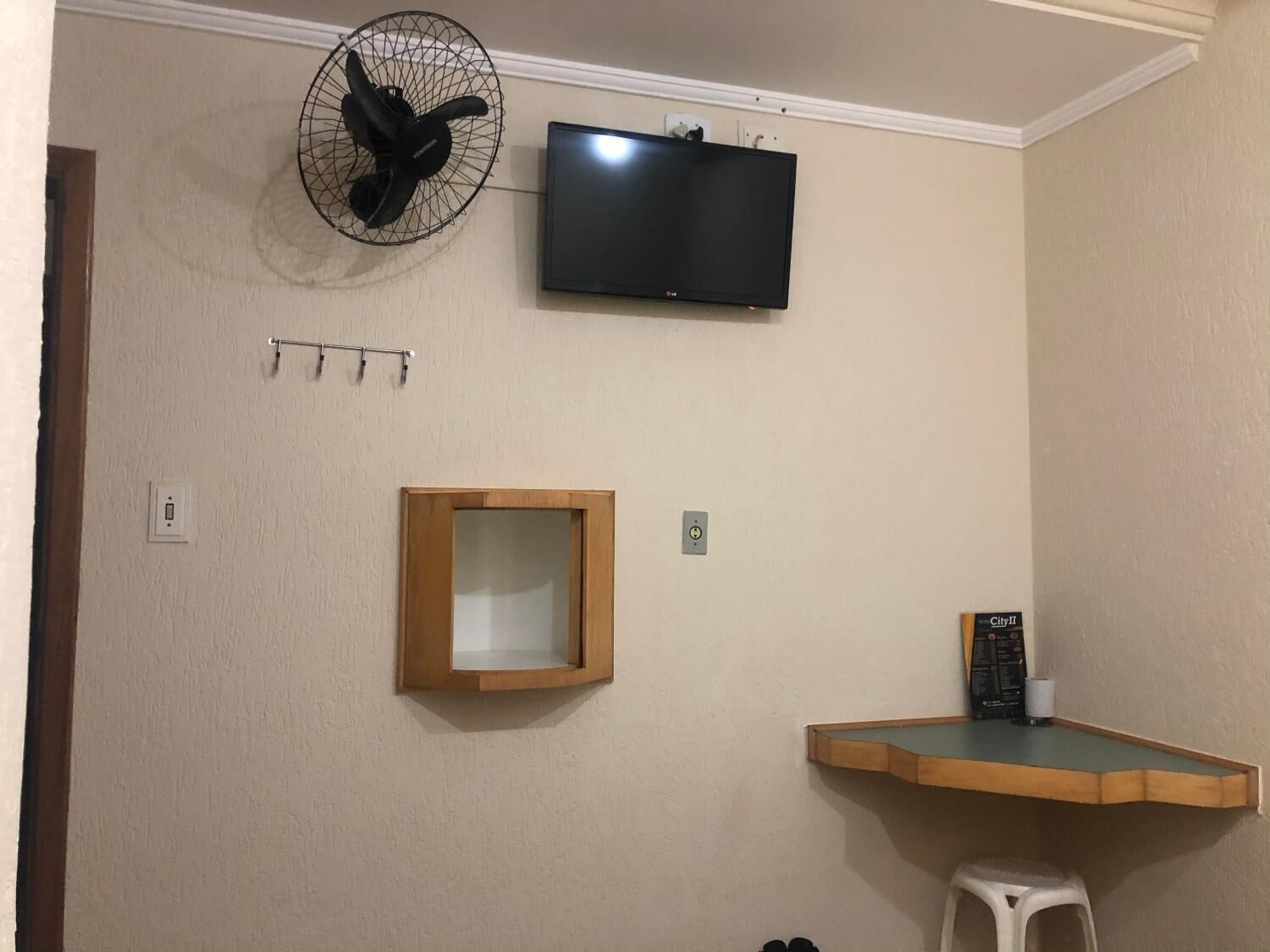 Quarto com televisão presa à parede, ventoinha fixada, prateleira de madeira com um pequeno banquinho e espelho emoldurado.