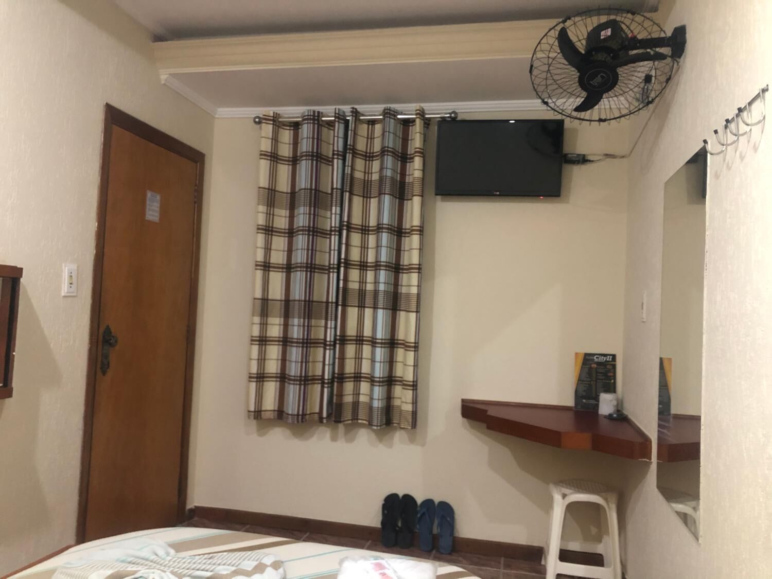 A room with a plaid curtain, a ceiling fan, a TV mounted on the wall, a wooden door, and a small shelf with items on it.
