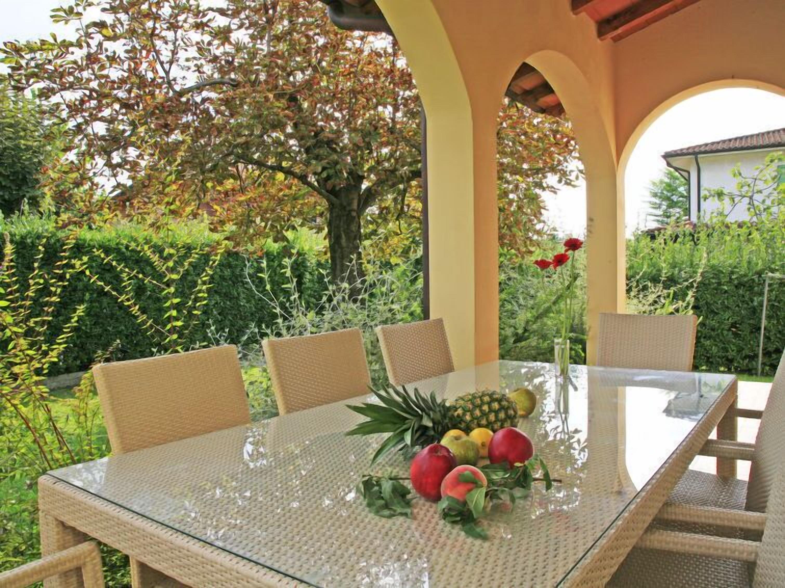 An outdoor dining area with a glass table surrounded by wicker chairs, set against a backdrop of lush greenery and a tree.