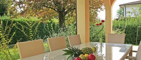 Un espacio exterior para comer con una mesa de vidrio rodeada de sillas de mimbre, con abundante follaje y un árbol de fondo.