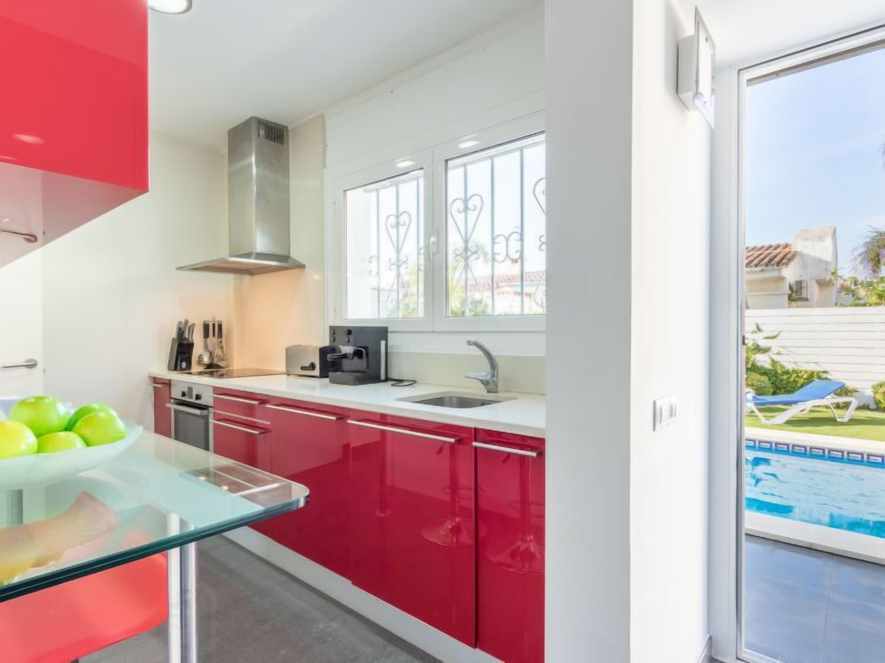 A modern kitchen with red cabinets, a glass countertop, and a view of a swimming pool.