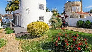 Ein weißes Gebäude mit geschwungener Fassade, ein kleiner Garten mit gelben und roten Blumen und ein geparktes Auto.