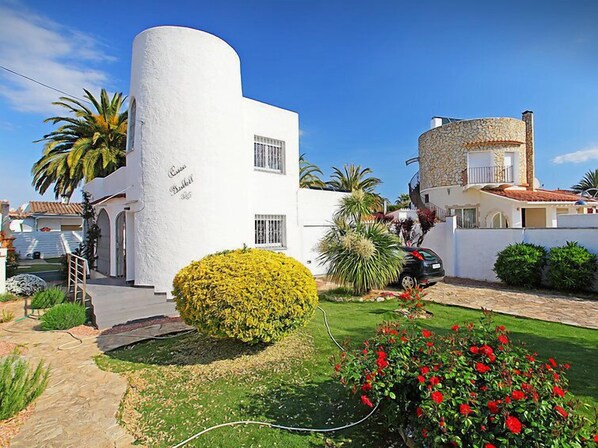 A white building with a curved facade, a small garden with yellow and red flowers, and a parked car.