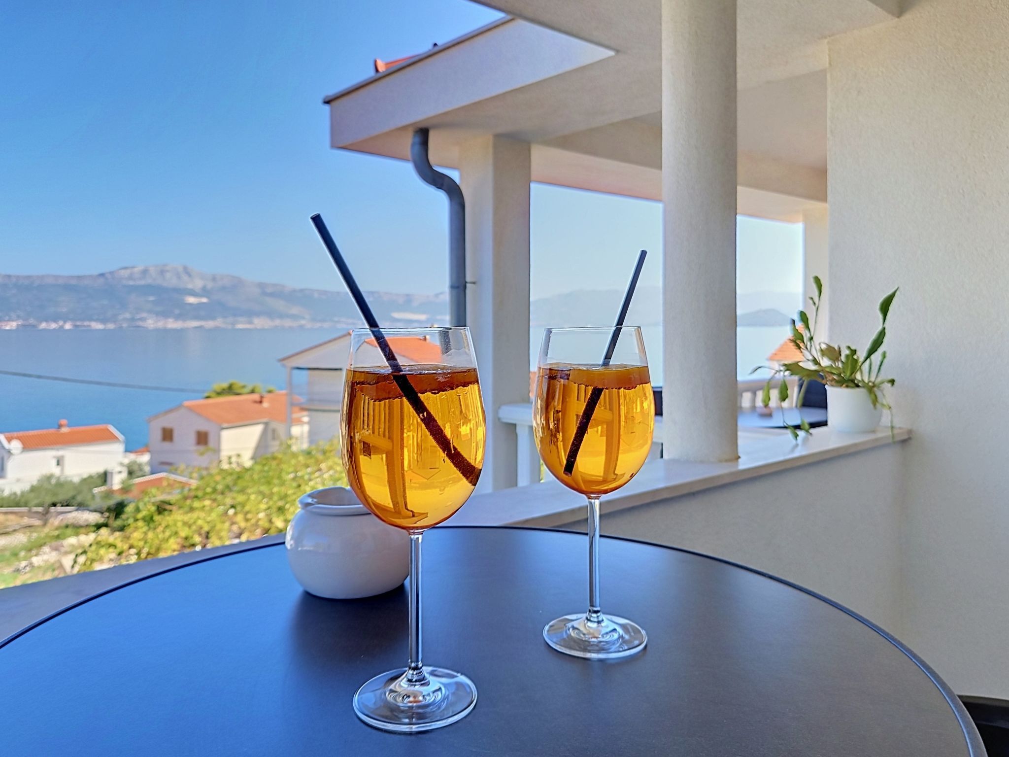 Two glasses of amber liquid with straws on a round table, overlooking a coastal landscape with buildings and mountains.