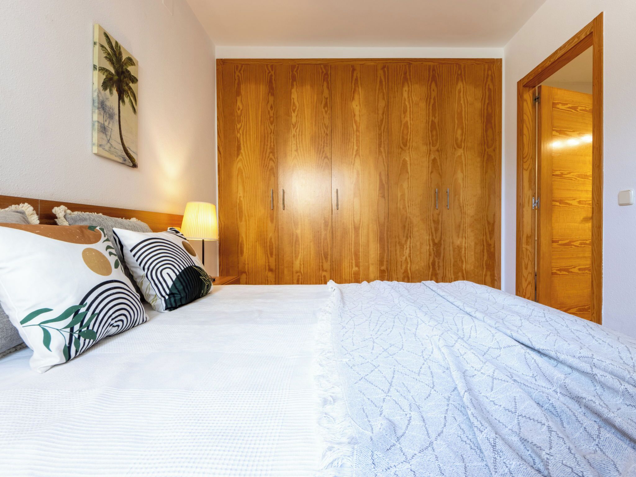 A bedroom with a wooden wardrobe, a bed with patterned bedding, a nightstand with a lamp, and a wall-mounted picture.