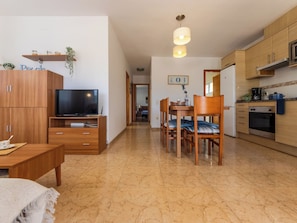 Un salon moderne avec un téléviseur, des meubles en bois, un coin repas avec des chaises, une cuisine avec des placards en bois et un couloir menant à une autre pièce.