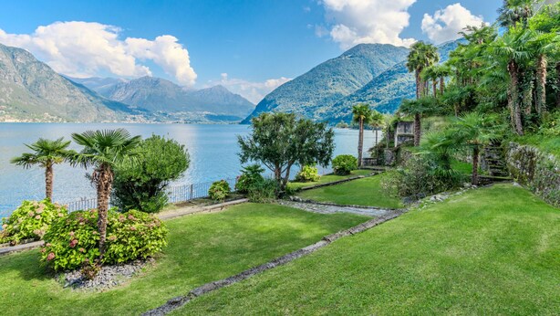 A lush garden with a pathway leading to a lake, surrounded by mountains and palm trees.