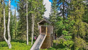 A wooden cabin with a wooden deck and a metal slide in a forested area.
