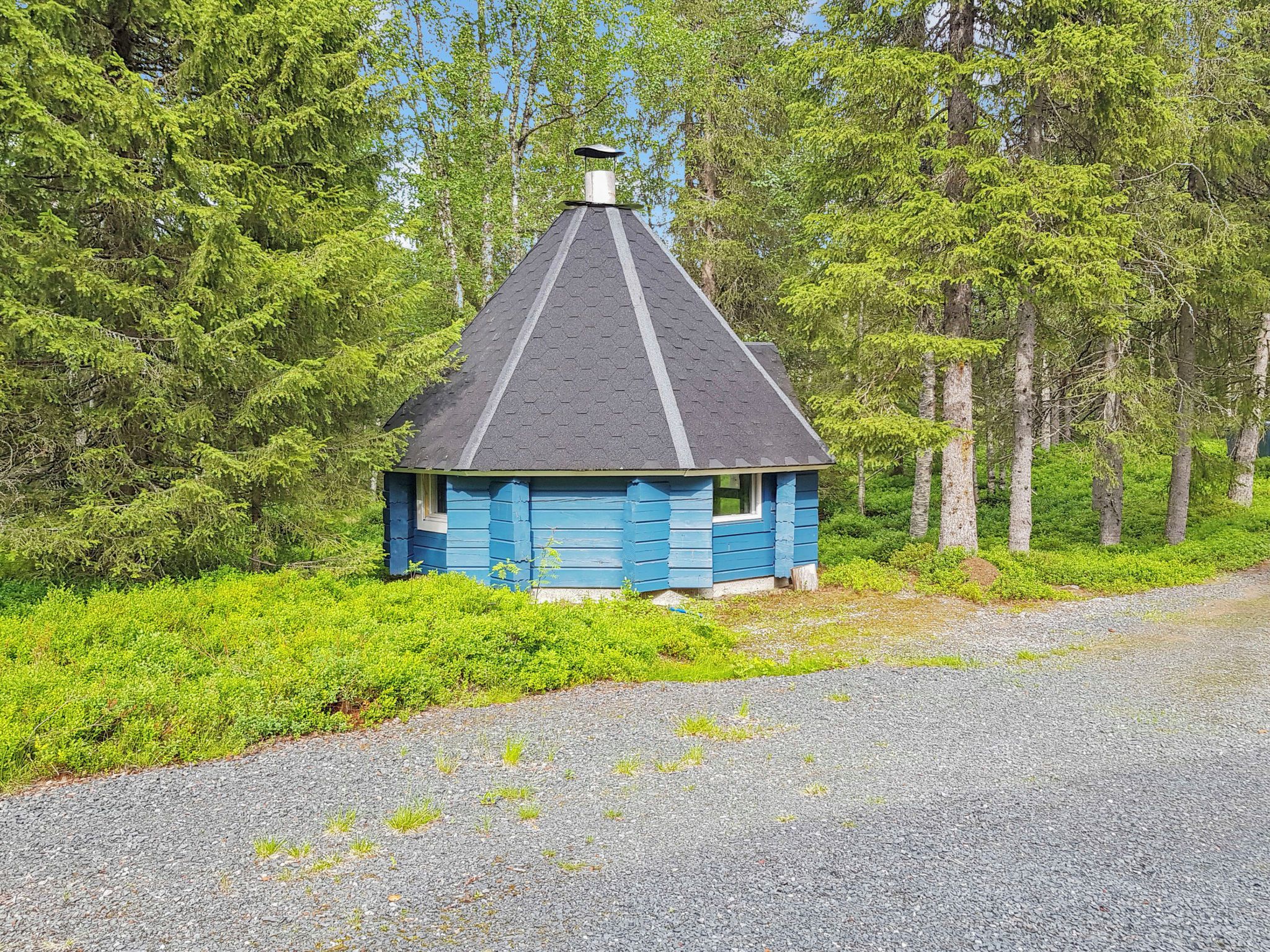 Een klein, blauw blokhutje met een puntdak, omgeven door weelderige groene begroeiing en bomen.