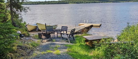 En campingplads ved søen med en træbro, et bord, stole og et bålsted.