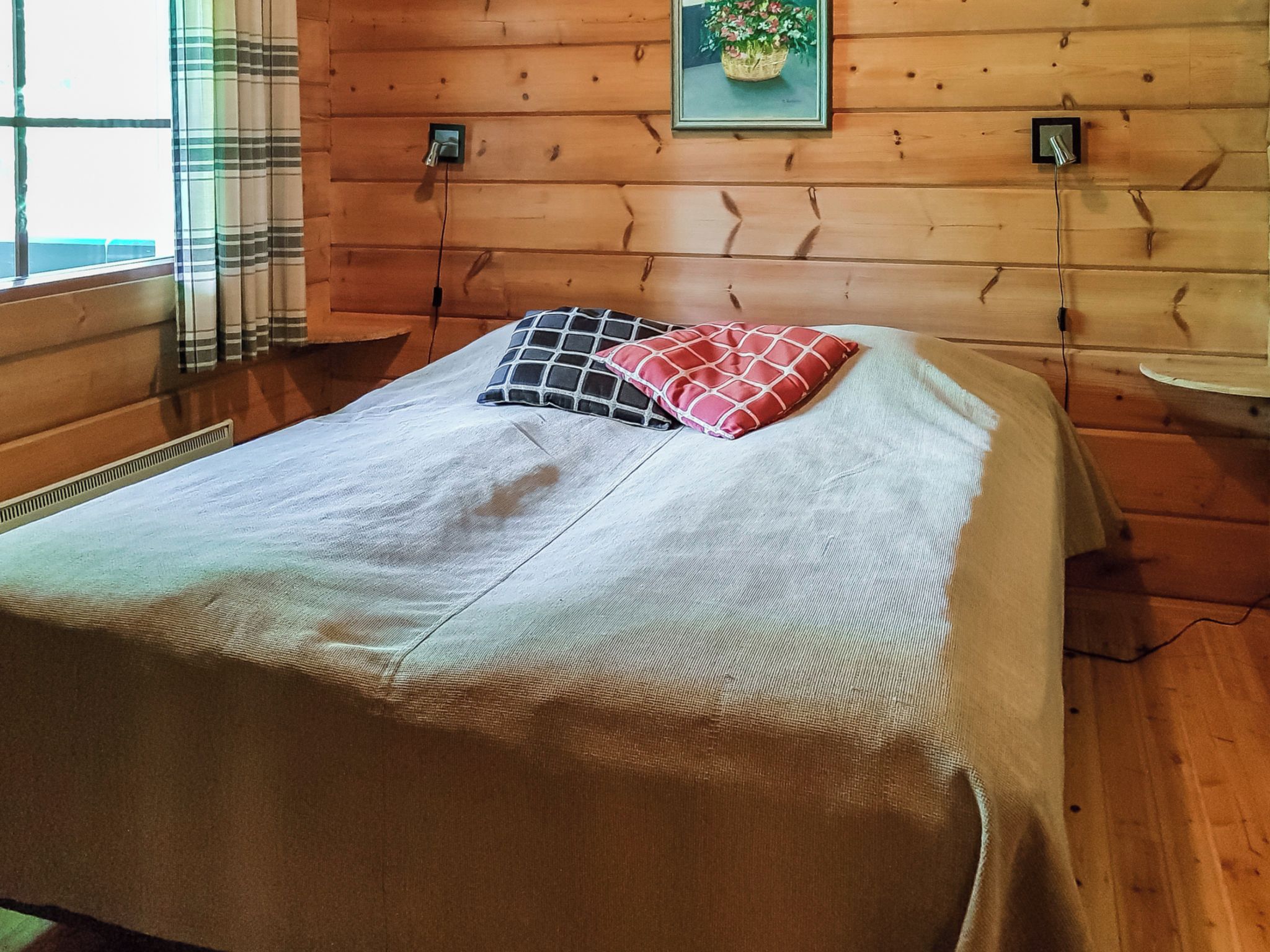 A bed with a white bedspread and two pillows, a window with curtains, and a wooden wall with a picture frame.