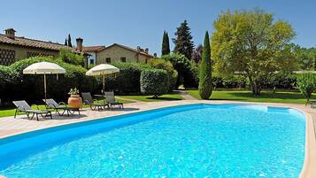 Una piscina con lettini e ombrelloni, circondata da siepi curate e un cielo sereno.