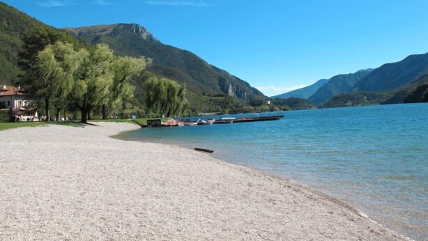Una spiaggia sul lago con una fila di barche, un edificio con tetto rosso e montagne sullo sfondo.