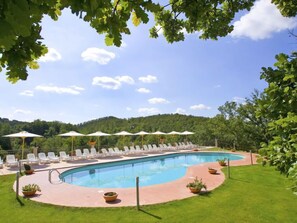 Ein Schwimmbecken mit Liegestühlen und Sonnenschirmen, umgeben von Grünflächen und Hügeln.