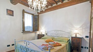 A bedroom with a metal bed frame, a chandelier, a window with blinds, and a bedside table with a lamp.