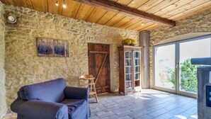 Una habitación rústica con pared de piedra, vigas de madera en el techo, un sofá azul, un mueble de madera y un ventanal con vistas a la vegetación.