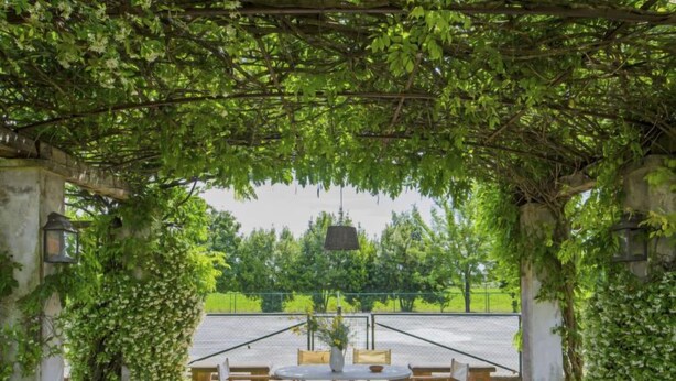 En uteplats med en pergola som är täckt av grönska, ett bord med stolar och en utsikt över en parkeringsplats och träd i bakgrunden.