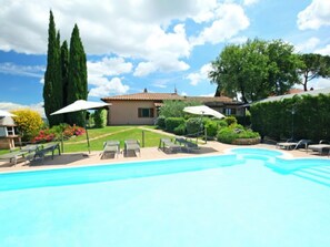 Une piscine entourée de chaises longues et de parasols, elle-même bordée d’un jardin bien entretenu et d’une maison dont le toit est recouvert de tuiles.