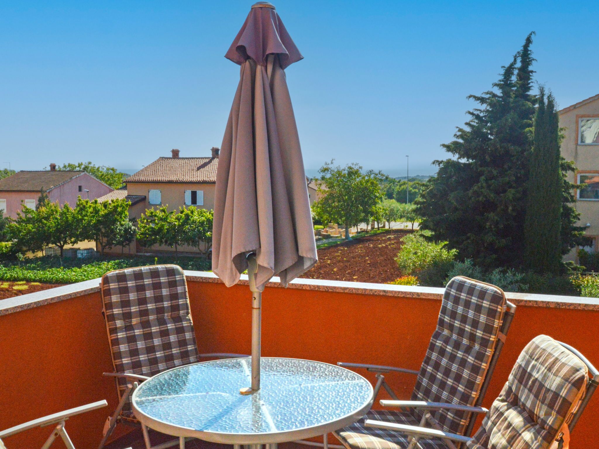 Un balcon avec une table ronde en verre et deux chaises à motifs écossais, un parasol et une vue sur des maisons et des arbres.