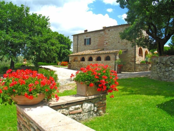Un edificio de piedra con techo de tejas, rodeado de abundante vegetación y flores rojas.