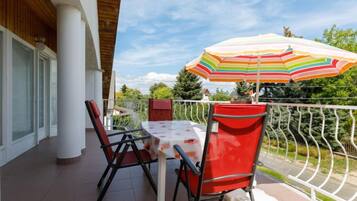A balcony with a table and chairs, a colorful striped umbrella, and a view of greenery and a building in the background.