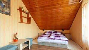 Een kleine, knusse slaapkamer onder het dak met een schuine houten plafond, een bed met een paars-wit geruit beddegoed, een klein tafeltje met een stoel, en een wandplank.