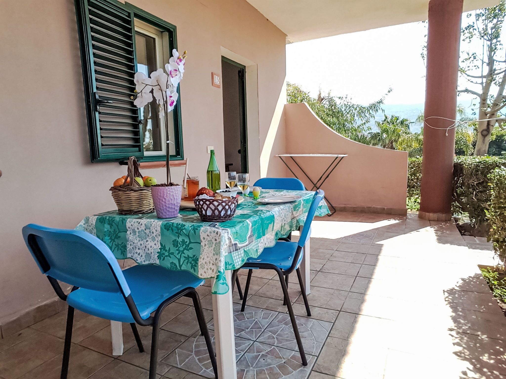 Mesa posta ao ar livre com cadeiras, toalha de mesa e diversos itens, incluindo frutas, garrafa e cesta.