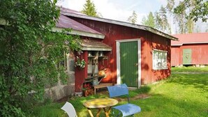 Un bâtiment en bois rouge avec une porte verte, un petit porche avec une table et des chaises, et une cour couverte d’herbe.