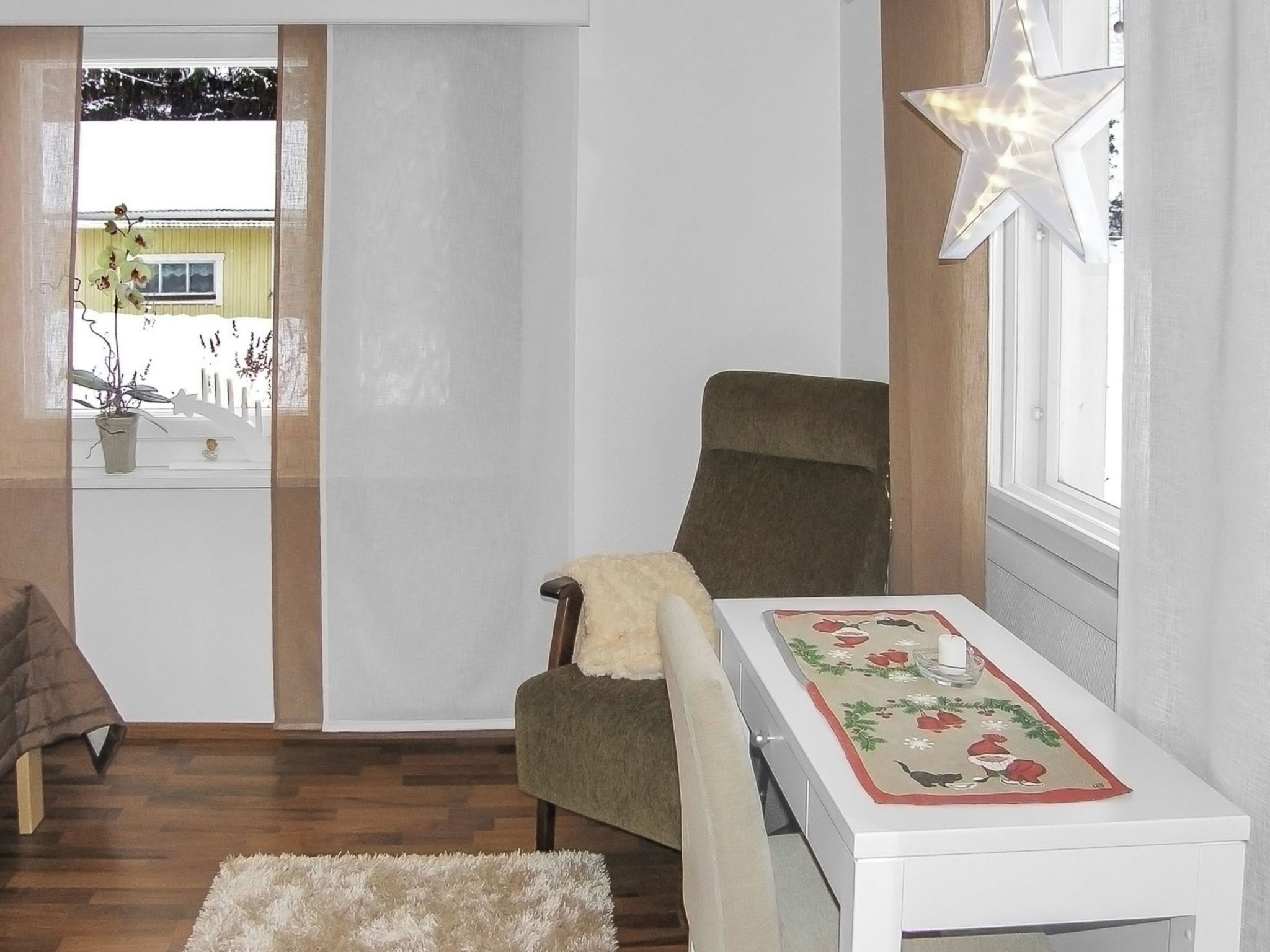 Ein Zimmer mit Holzboden, einem grünen Sessel, einem weißen Tisch mit floralem Tischtuch, einer sternförmigen Leuchte und Blick auf ein Haus durch ein Fenster.