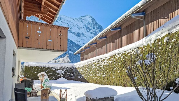 A snow-covered outdoor area with a wooden building, a table with chairs, and a potted plant. - Chalet am Reeti by Interhome (Grindelwald)