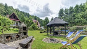 A stone-built outdoor kitchen with a fire pit, wooden benches, and lounge chairs in a lush garden setting. - Vacation home Hatale by Interhome (Rzyki)