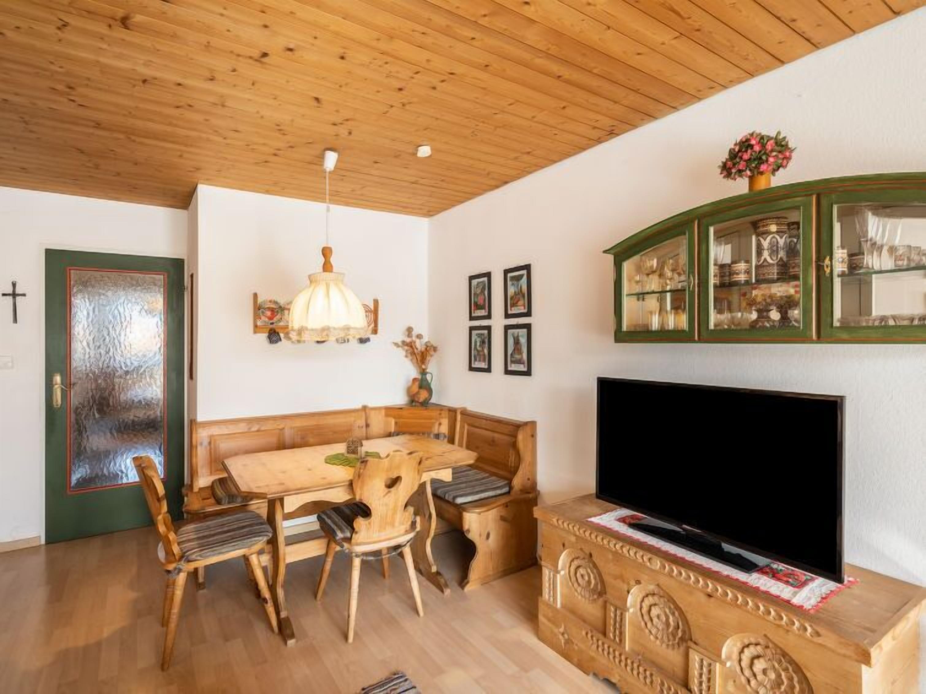 Ein gemütliches Wohnzimmer mit Holzbalkendecke, einem Esstisch mit Stühlen, einer Couch, einem Fernseher auf einem Holzschrank und einem grünen Schrank mit Glastüren.