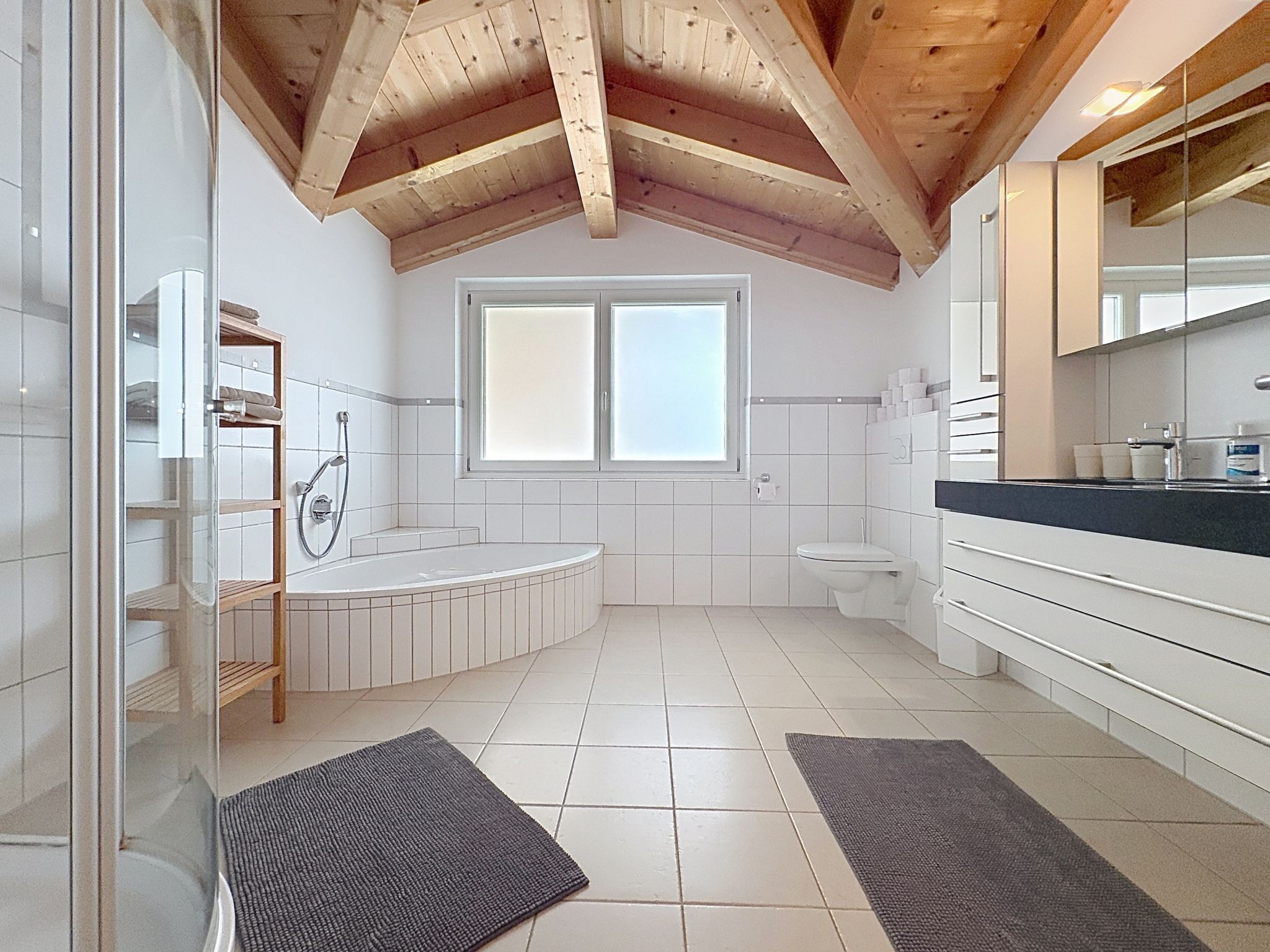 Ein modernes Badezimmer mit Badewanne, Toilette und Dusche, Holzbalkendecke und einem großen Fenster.